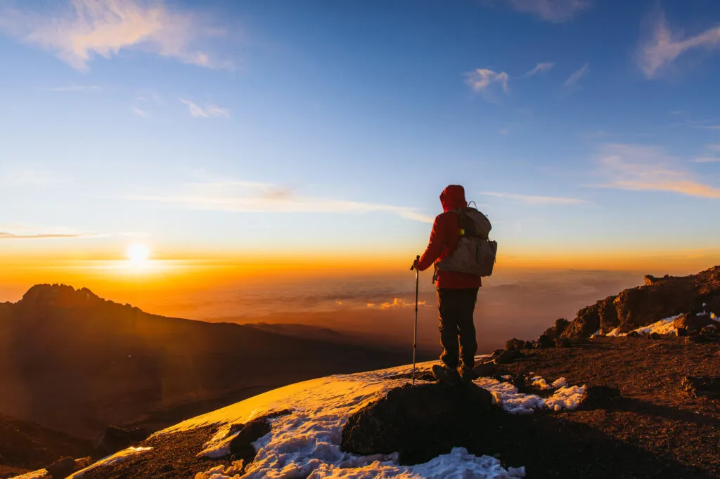 Silhouet van een bergbeklimmer met rugzak en wandelstokken die op een sneeuwachtige bergtop staat en kijkt naar een spectaculaire zonsopgang boven het wolkendek op Mount Kilimanjaro in Tanzania.