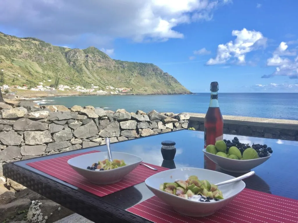Een gedekte eettafel met kommetjes eten en een fles drank op de voorgrond, met uitzicht op een blauwe baai en groene bergen bij Vigia da Areia op Santa Maria in de Azoren.