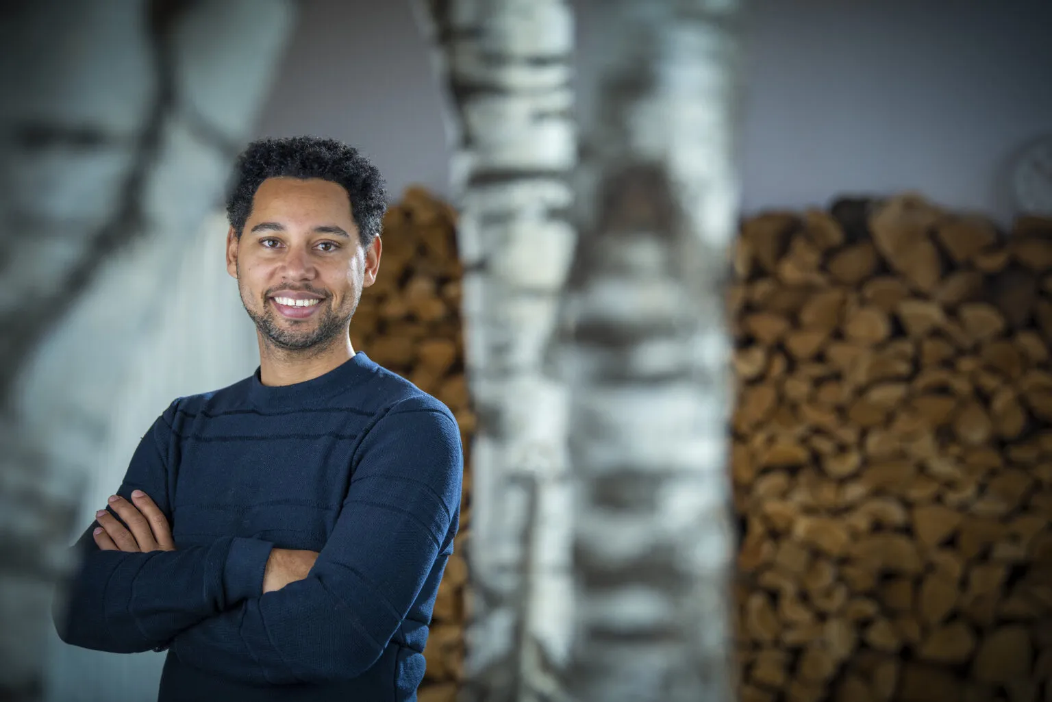 Portret van Joshua van Eijndhoven, oprichter van Voja Travel, glimlachend met gehaspelde armen voor een stapel brandhout.