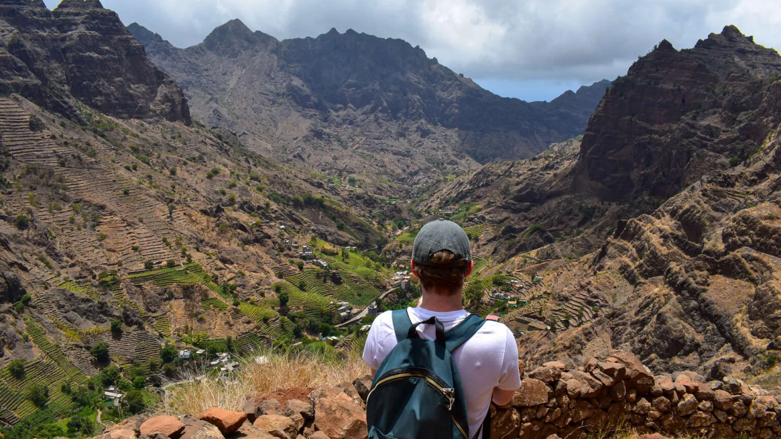 Rugweergave van een wandelaar met pet en rugzak die uitkijkt over een diepe bergvallei bij Caibros op Santo Antão, Kaapverdië.