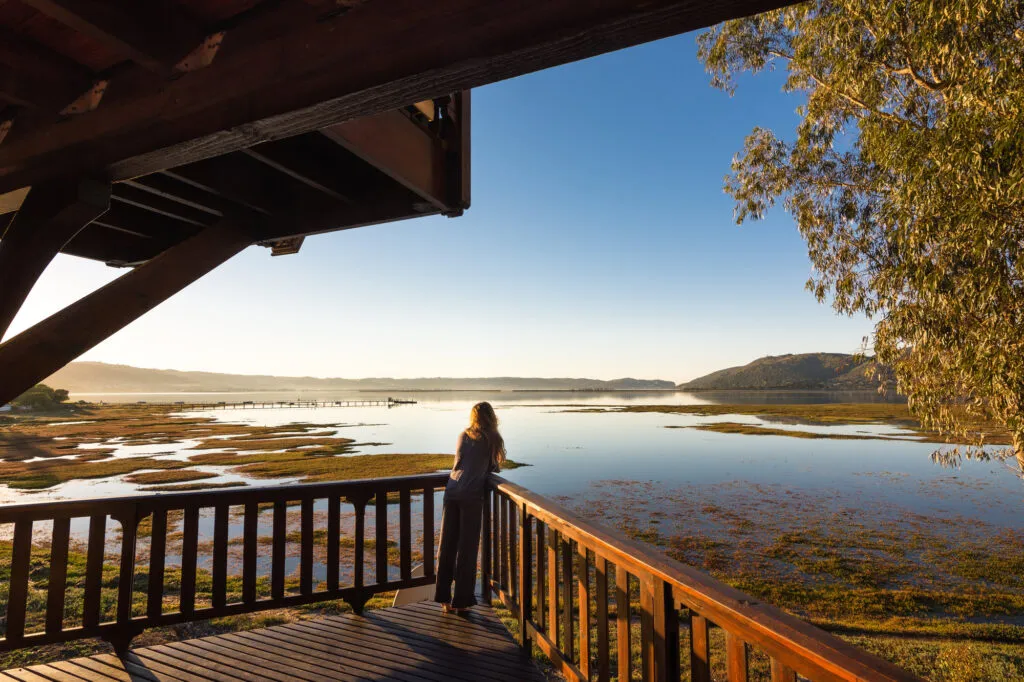 Aanzicht vanaf de rug van een vrouw die vanaf een houten balkon uitkijkt over het rustige water van de Knysna-lagune langs de Garden Route.