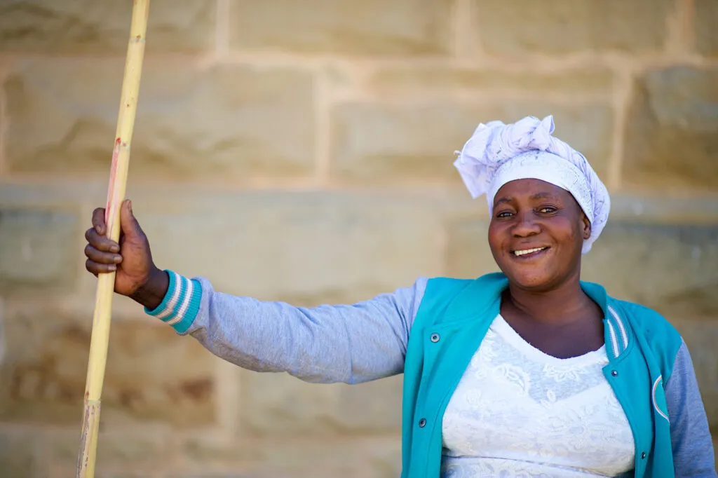 Portret van een lachende Zuid-Afrikaanse vrouw met een witte hoofddoek en een blauwe vest, staand voor een stenen muur in Oudtshoorn.