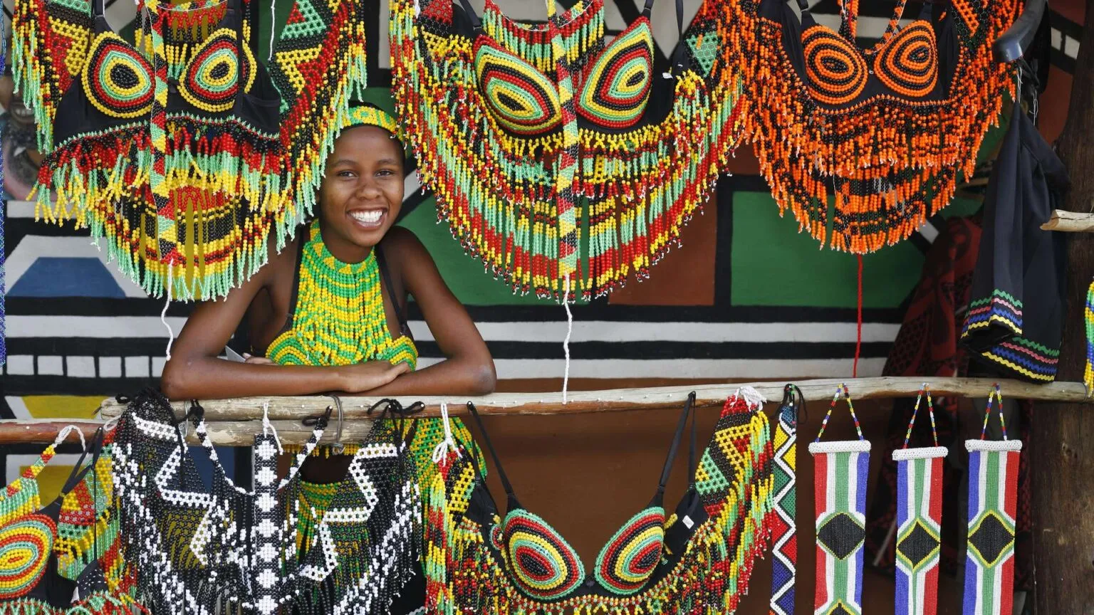 Portret van een glimlachende Zuid-Afrikaanse Zulu-vrouw die achter een balie staat omringd door kleurrijke, handgemaakte krallensieraden en traditioneel kraalwerk.