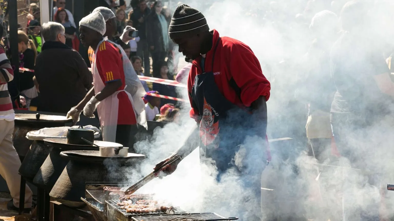 Zijaanzicht van een man met een rode jas en muts die vlees bereidt boven een rokende barbecue (braai) op een straatmarkt in Zuid-Afrika.