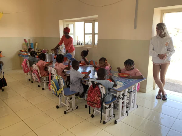Angelique staat in een klaslokaal waar een groep jonge kinderen aan kleine tafeltjes zit te eten, begeleid door een docente.