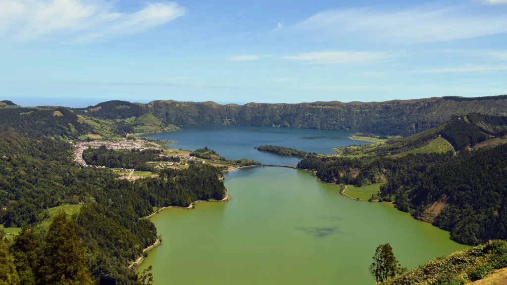 Panoramisch overzicht van het groene en blauwe kratermeer van Sete Cidades, gescheiden door een brug en omringd door steile groene hellingen.