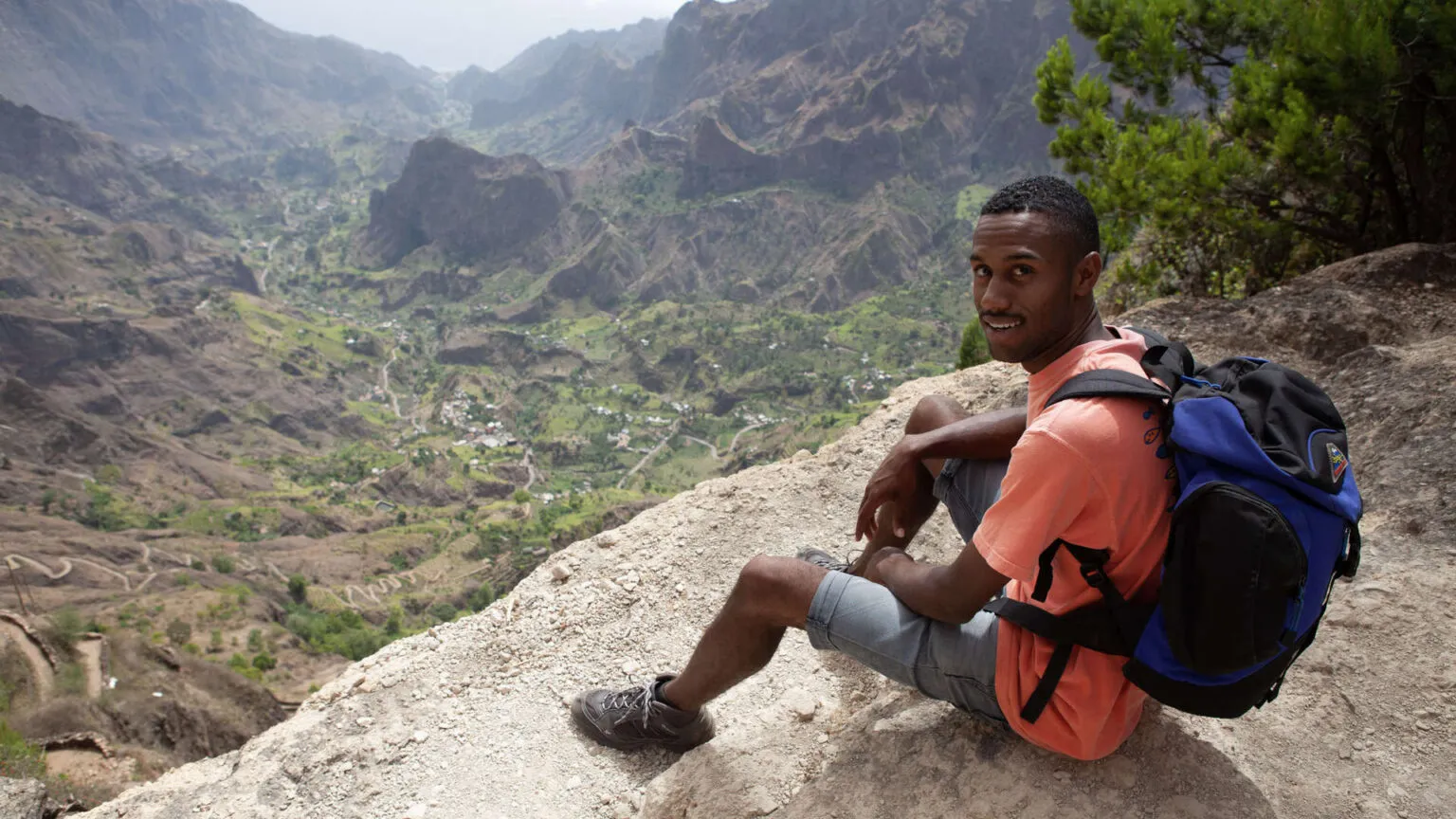 Gids Edson zit op een rotswand met een panoramisch uitzicht over de bergen van Santo Antão in Kaapverdië op de achtergrond.