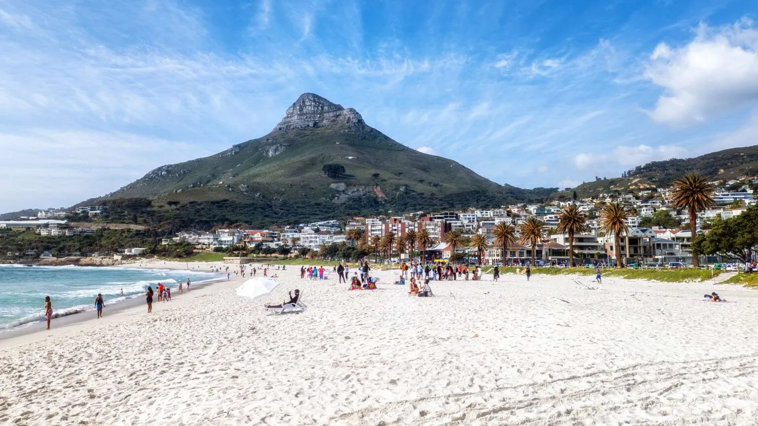 Zonnig overzicht van het witte zandstrand van Camps Bay in Kaapstad, gevuld met strandgangers, palmbomen en de iconische berg Lion's Head op de achtergrond.