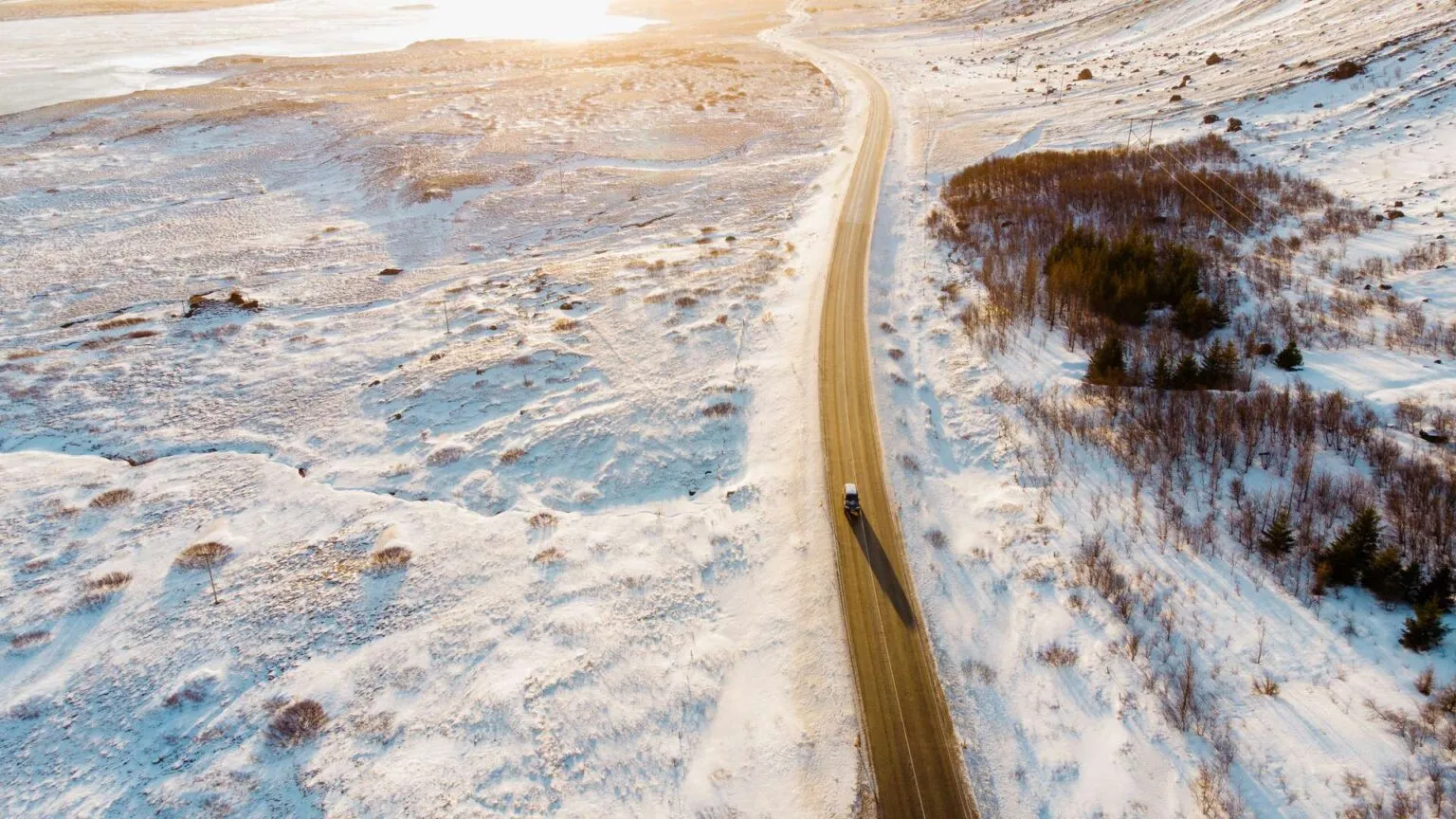 Bovenaanzicht van een asfaltweg die door een winters IJslands landschap meandert, omgeven door met sneeuw bedekte velden en lage vegetatie bij zacht zonlicht.