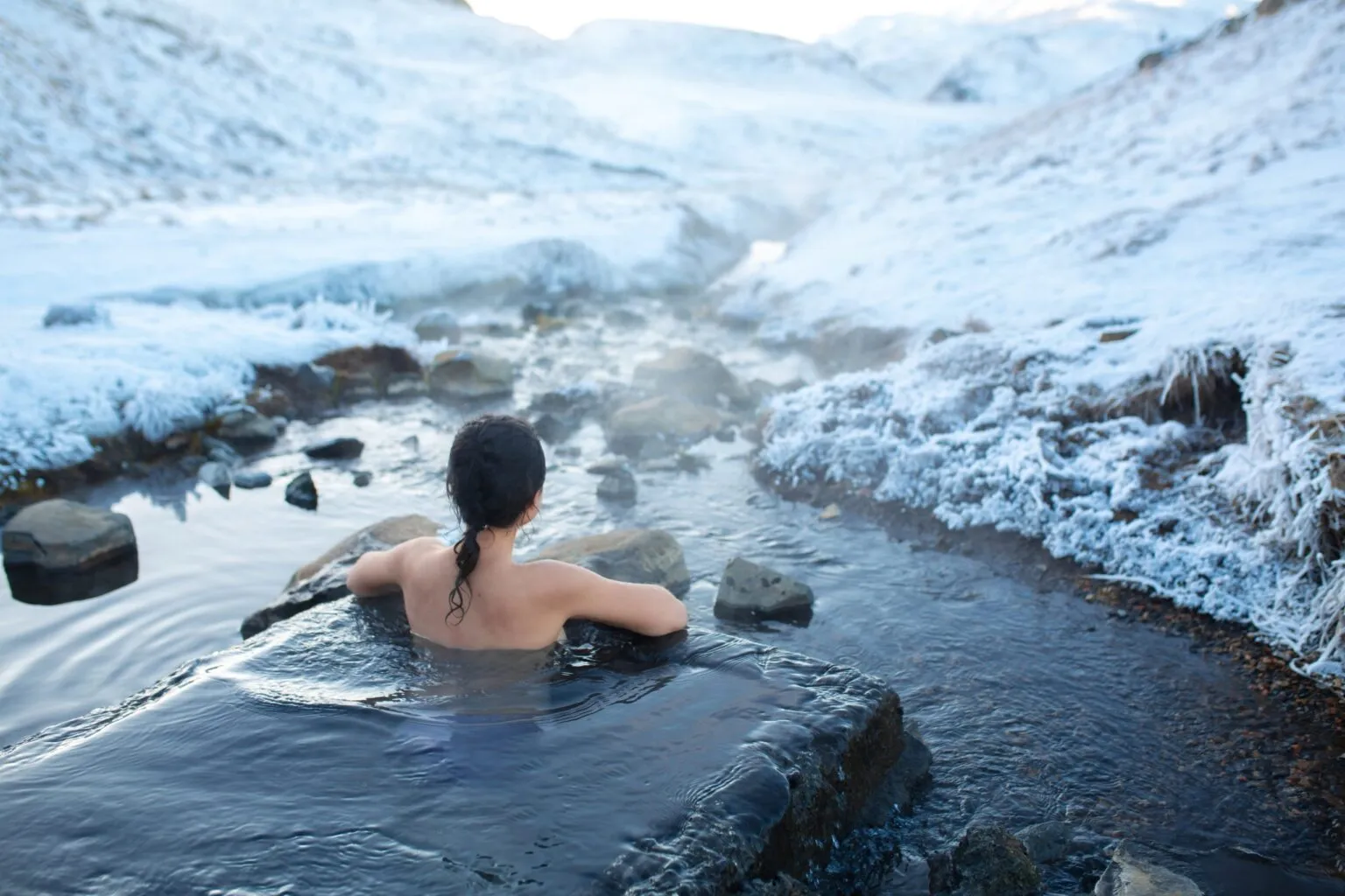 Een persoon zit van de achterkant gezien in een stomende natuurlijke hot spring midden in een met sneeuw bedekt IJslands berglandschap.