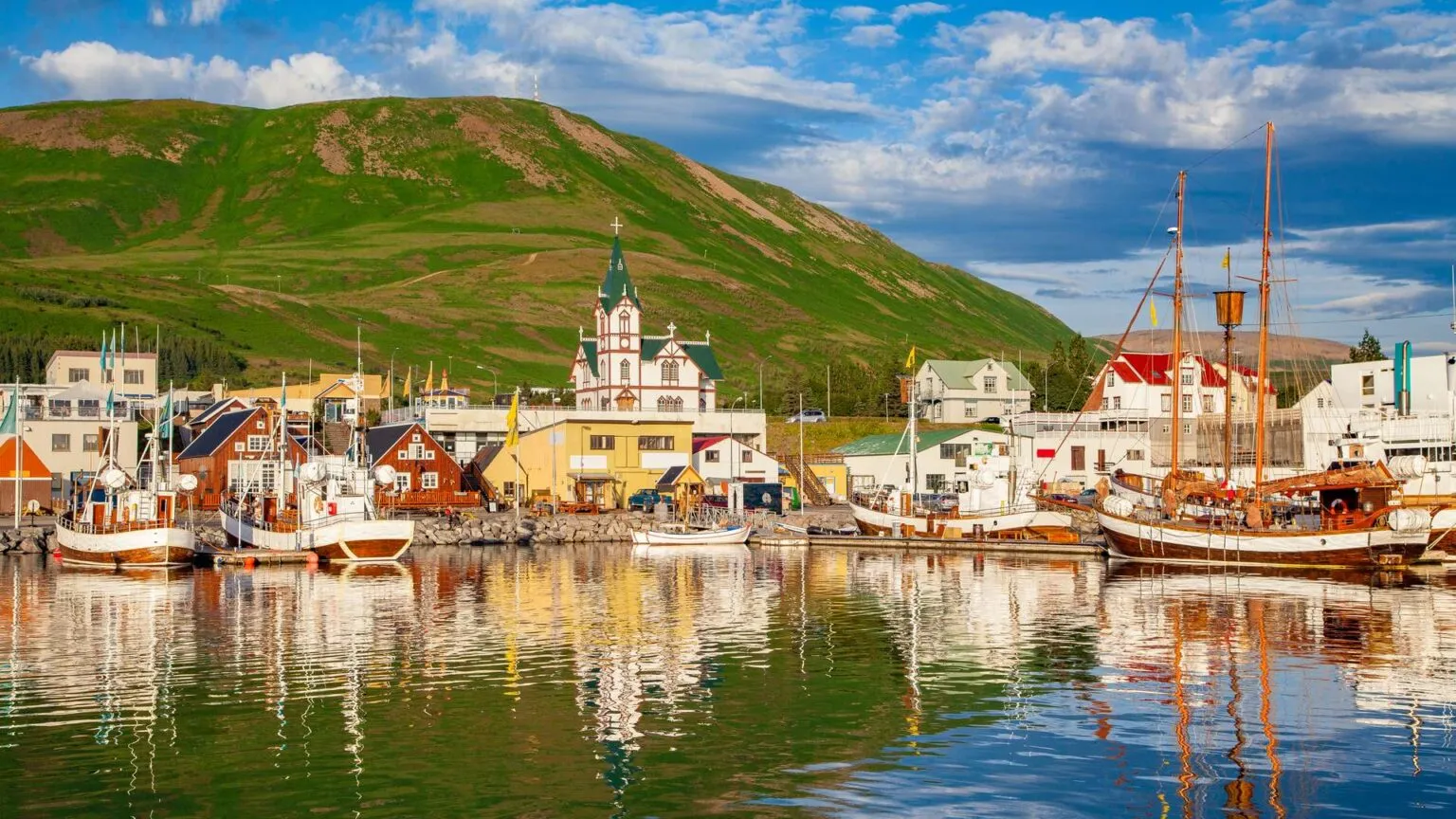 Kleurrijke houten huizen, de karakteristieke kerk en houten walvisspotboten weerspiegelen in het kalme water van de haven van Húsavík in IJsland.