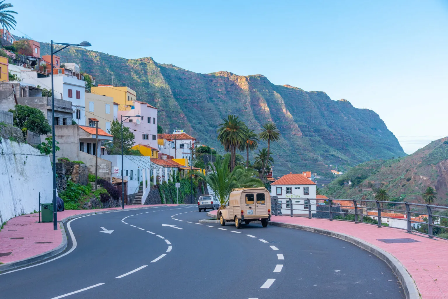 Een strakke asfaltweg kronkelt langs gekleurde huizen tegen een achtergrond van steile, groene bergen op La Gomera.