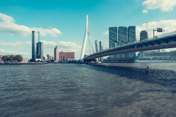 Zicht vanaf het water op de Erasmusbrug en de moderne skyline van Rotterdam onder een blauwe lucht met lichte bewolking.