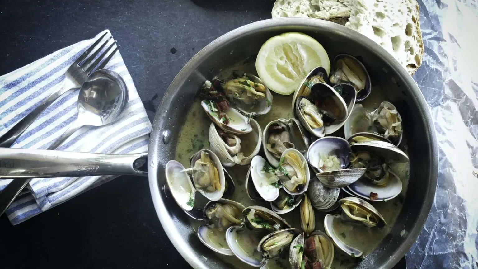 Een metalen pan gevuld met bereide schelpdieren in saus en een schijfje citroen, geserveerd met brood en bestek op Madeira.