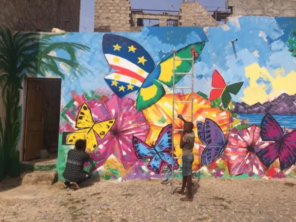 Twee personen werken aan een kleurrijke muurschildering met vlinders, waaronder een vlinder met de Kaapverdische vlag, in Mindelo op São Vicente.