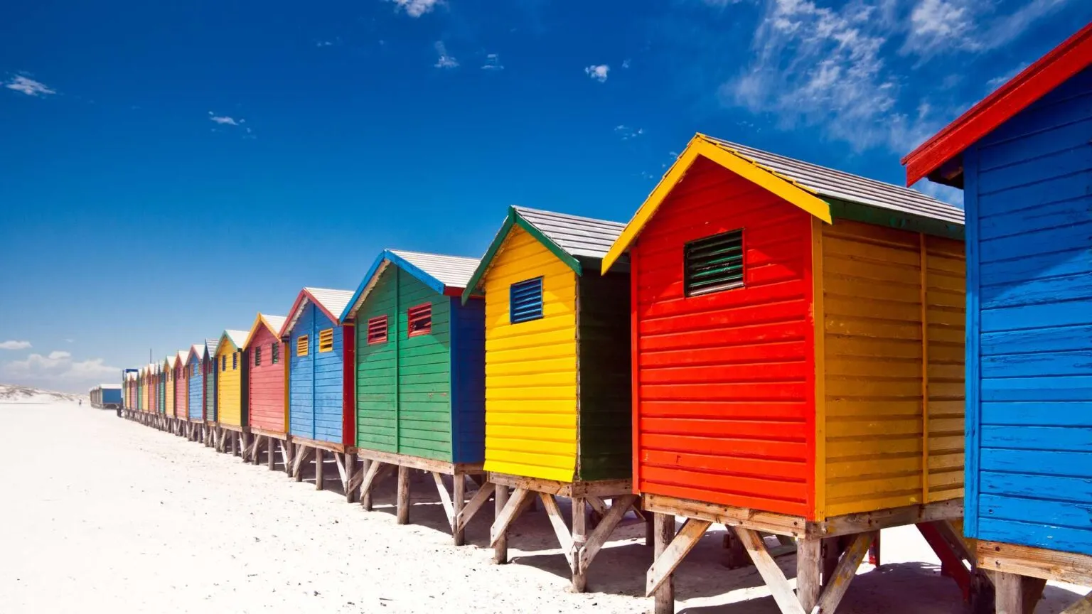 Rij van iconische, felgekleurde houten strandhuisjes in het geel, rood, groen en blauw op het zandstrand van Muizenberg onder een strakblauwe lucht.