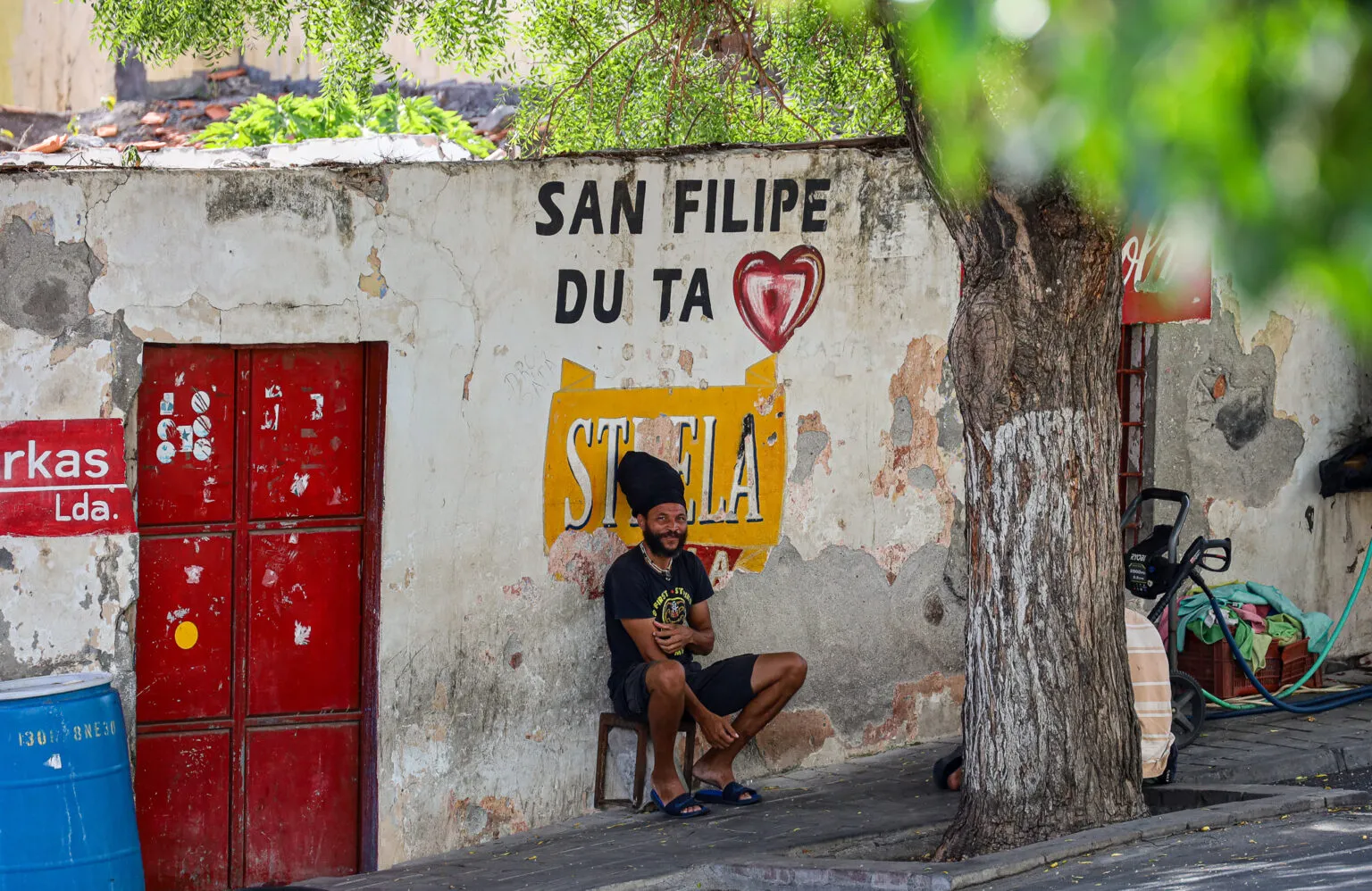 Een lokale man zit op een stoel op de stoep in São Filipe, Fogo; op de muur achter hem staat een schildering met 'San Filipe du ta' en een hartje