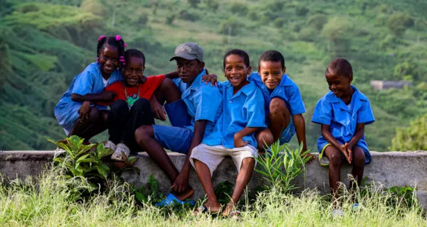 Een groep vrolijke schoolkinderen in blauwe schooluniformen zit samen op een muurtje in het groene berglandschap van Santiago, Kaapverdië.