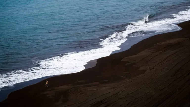 Een wandelaar op het vulkanische strand bij Sãõ Filipe op Fogo; het zwarte strand contrasteert sterk met de blauwe zee