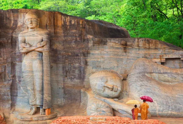 Een staande en een liggende Boeddha uitgehouwen in een massieve rotswand, met twee bezoekers met een paraplu op de voorgrond bij de Gal Vihara tempel.
