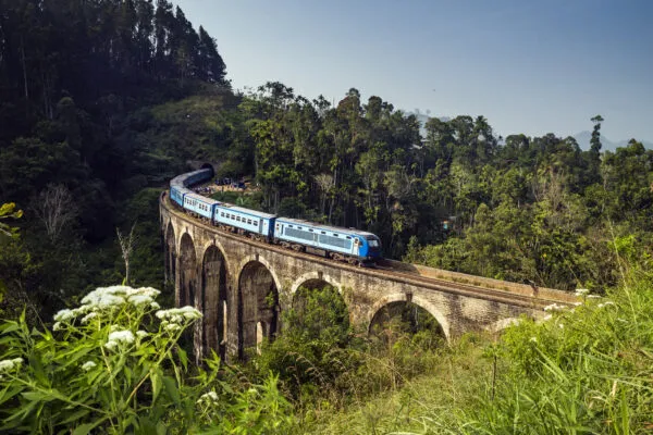 Een blauwe passagierstrein rijdt over een stenen stenen viaduct, omgeven door dicht groen bos en tropische vegetatie in de bergen van Sri Lanka.