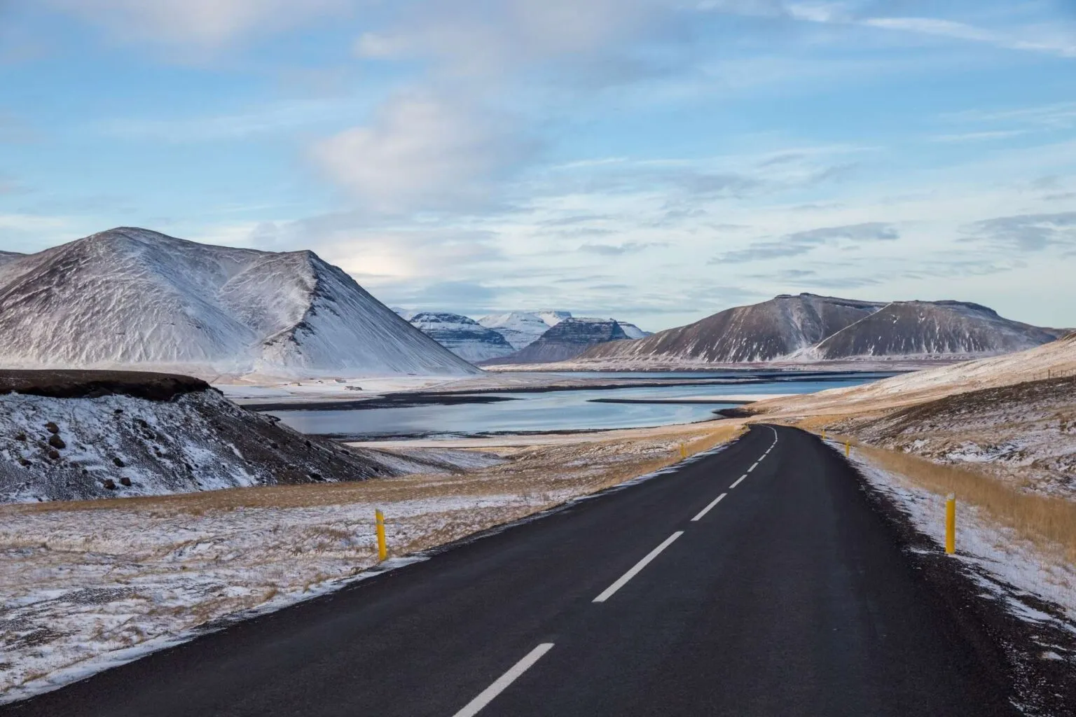 Een zwarte asfaltweg meandert door een winters landschap met sneeuwbedekte bergen en een meer of fjord onder een heldere lucht in IJsland.