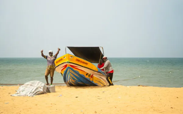 Een man in een bloemig shirt staat op het zandstrand van Negombo en zwaait met beide handen naar de camera, naast een kleurrijk beschilderde vissersboot.