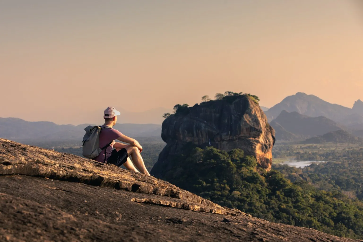 Een wandelaar met een rode rugzak zit op de top van een berg en kijkt uit over de iconische Sigiriya Lion Rock in Sri Lanka tijdens een goudkleurige zonsondergang.