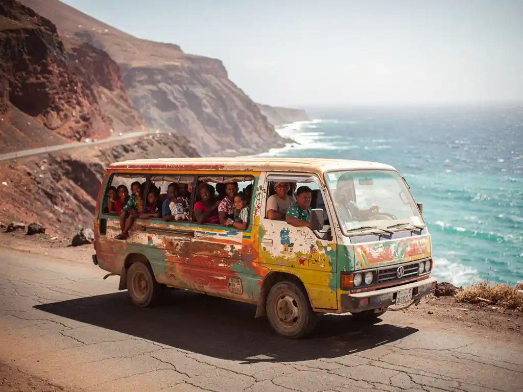 Kleurrijke aluguer-minibus vol passagiers op een slingerende kustweg in Kaapverdië, met vulkanische kliffen en turquoise Atlantische Oceaan.