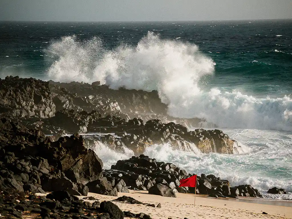 Krachtige oceaangolf die breekt op het vulkanische basaltrotskust van Kaapverdië, met wit schuim en een waarschuwingsvlag op het strand.