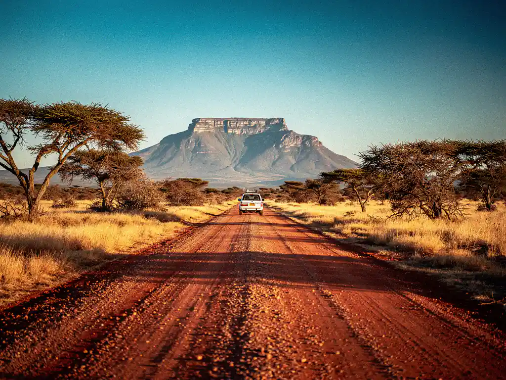 Witte 4x4 huurauto op rood grindpad door Zuid-Afrikaanse savanne, met tafelvormige berg in de verte tijdens gouden uur.