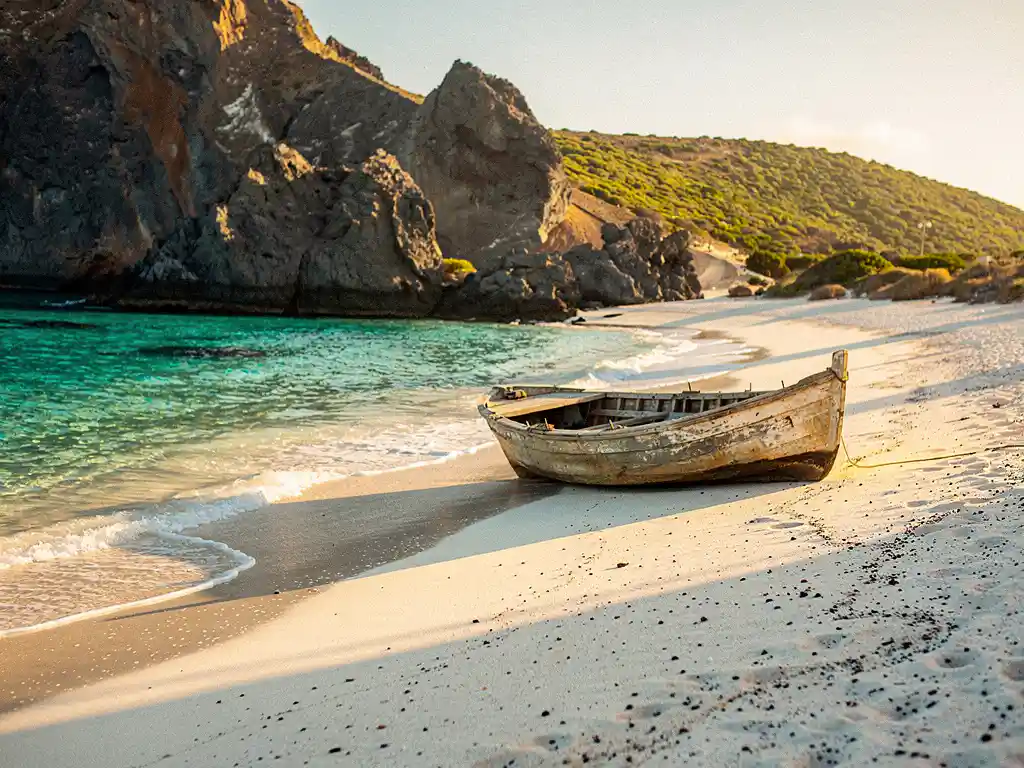 Verouderde houten vissersboot op wit vulkanisch zand aan een turquoise baai in Kaapverdië, met dramatische kliffen op de achtergrond.