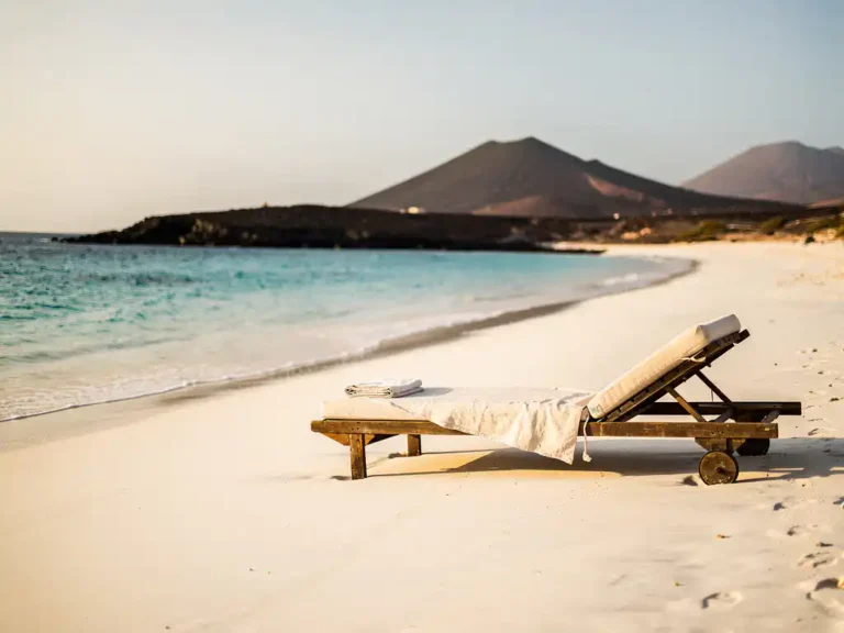 Wit zandstrand op Kaapverdië met turquoise water, een houten ligstoel met linnen handdoek en vulkanische heuvels op de achtergrond.