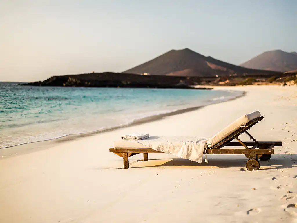 Wit zandstrand op Kaapverdië met turquoise water, een houten ligstoel met linnen handdoek en vulkanische heuvels op de achtergrond.
