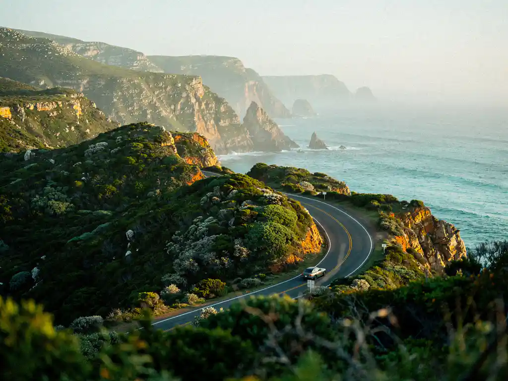 Kronkelende kustweg door groene fynbos boven de turquoise Indische Oceaan, met mistige rotsachtige kapen op de achtergrond en één auto in de bocht.