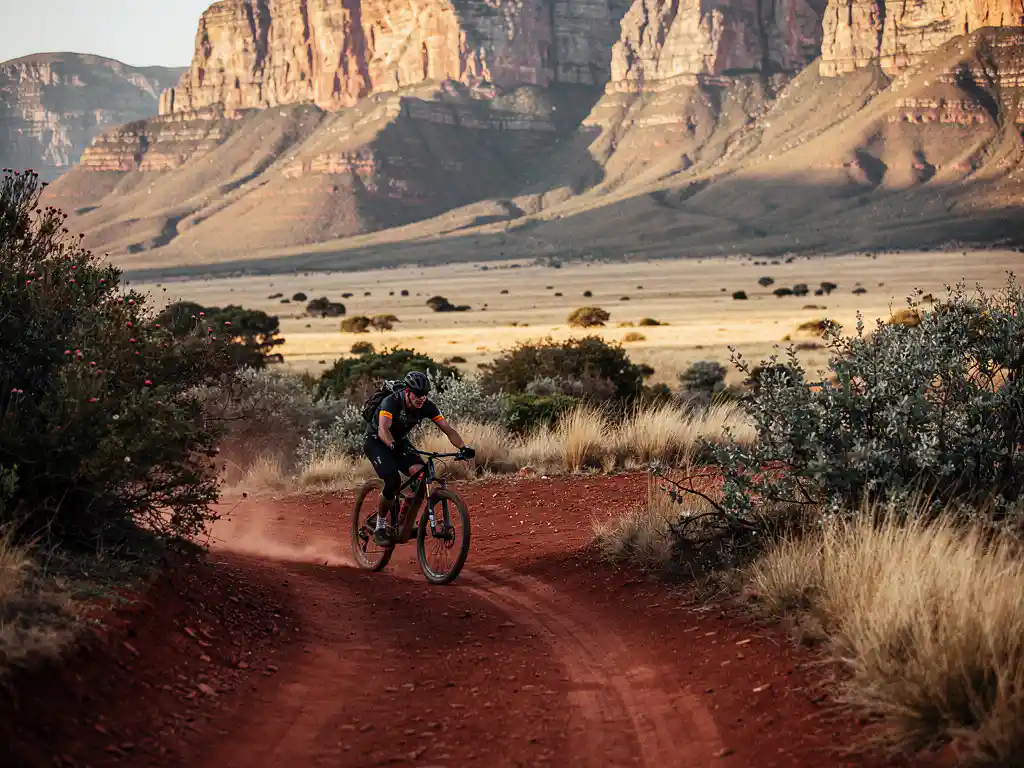Mountainbiker op rood stofpad langs de Drakensbergkliffen in Zuid-Afrika, omgeven door fynbos en gouden savanne in middaglicht.