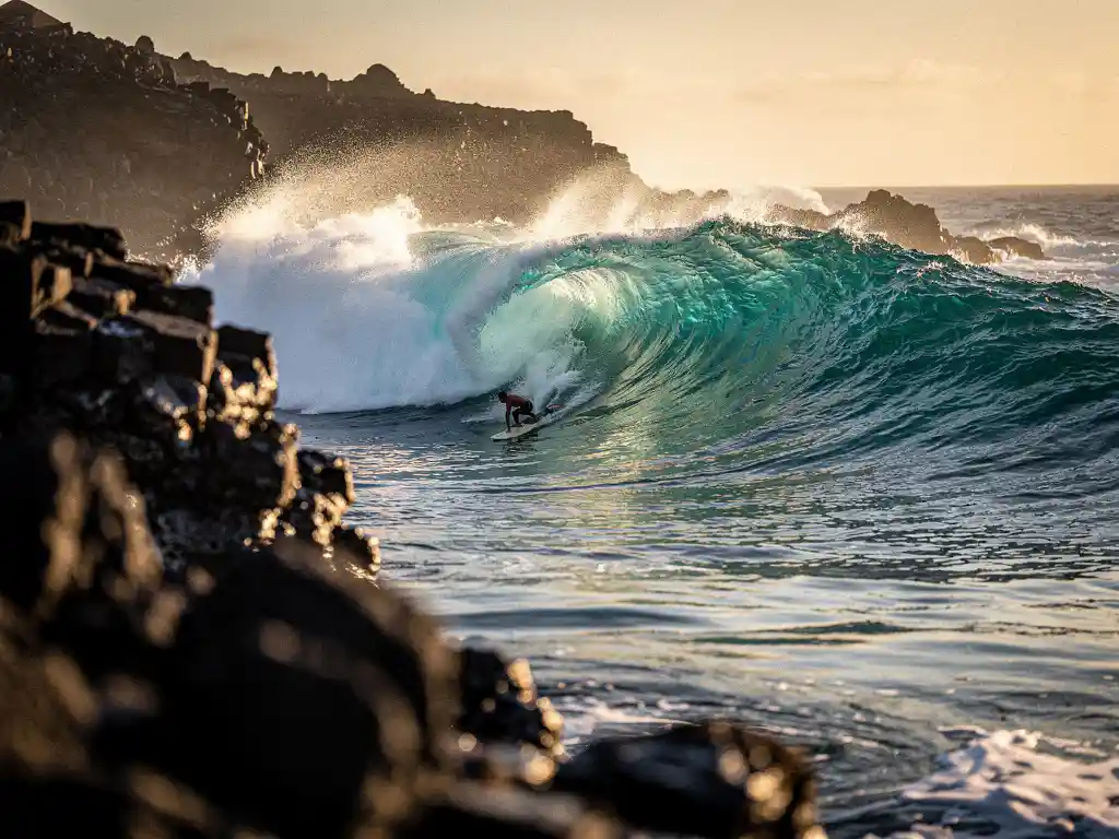 Surfer peddelt richting krachtige Atlantische golf bij Cape Verde, omgeven door donkere basaltrotsen en turquoise water in middagzon.