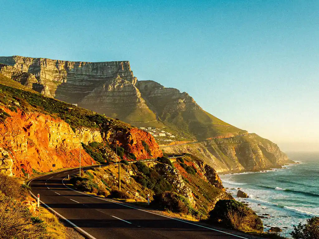 Tafelberg met plat silhouet boven groene hellingen in Kaapstad, kronkelende kustweg naar turquoise Atlantische Oceaan in gouduur licht.