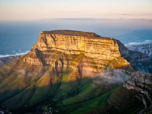 Luchtfoto van de vlakke top van de Tafelberg boven Kaapstad, met groene hellingen, gouden ochtendlicht en de Atlantische Oceaan.
