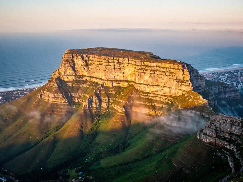 Luchtfoto van de vlakke top van de Tafelberg boven Kaapstad, met groene hellingen, gouden ochtendlicht en de Atlantische Oceaan.