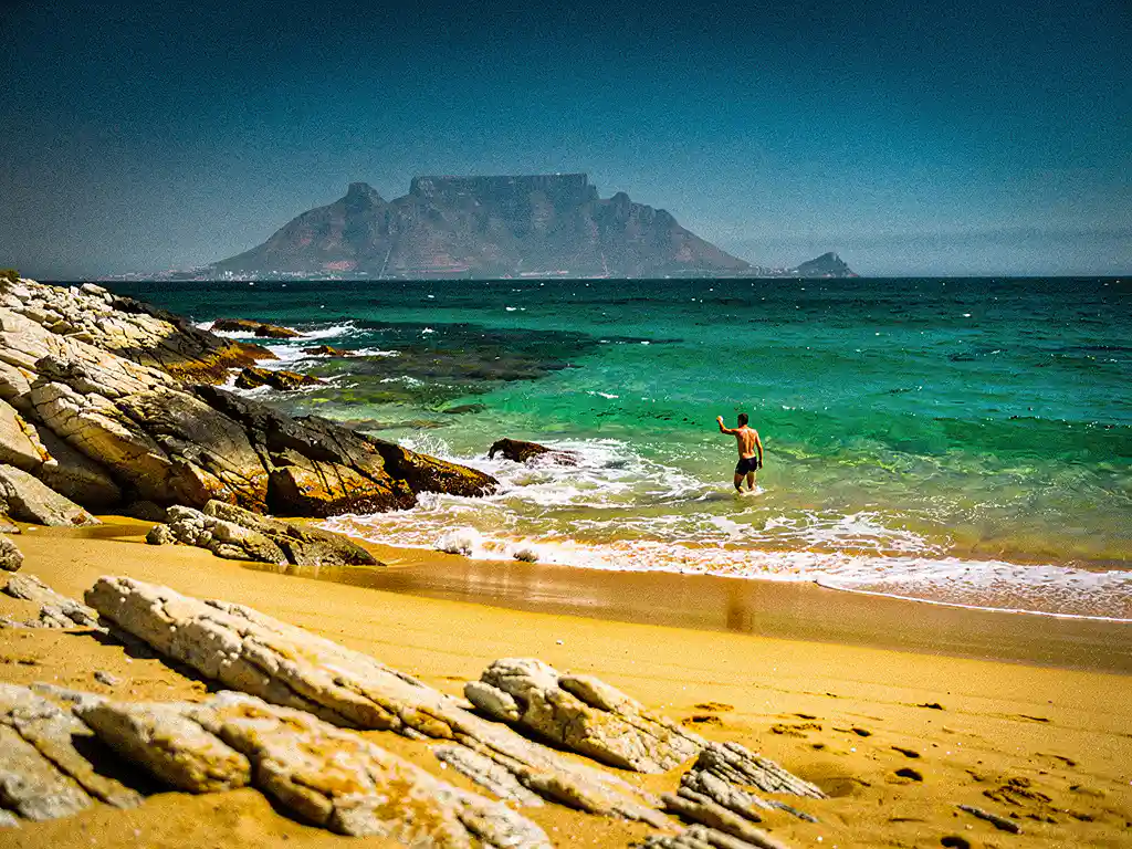 Zwemmer die de turquoise Atlantische Oceaan inloopt bij een gouden zandbaai aan de Zuid-Afrikaanse kust, met de Tafelberg in de verte.