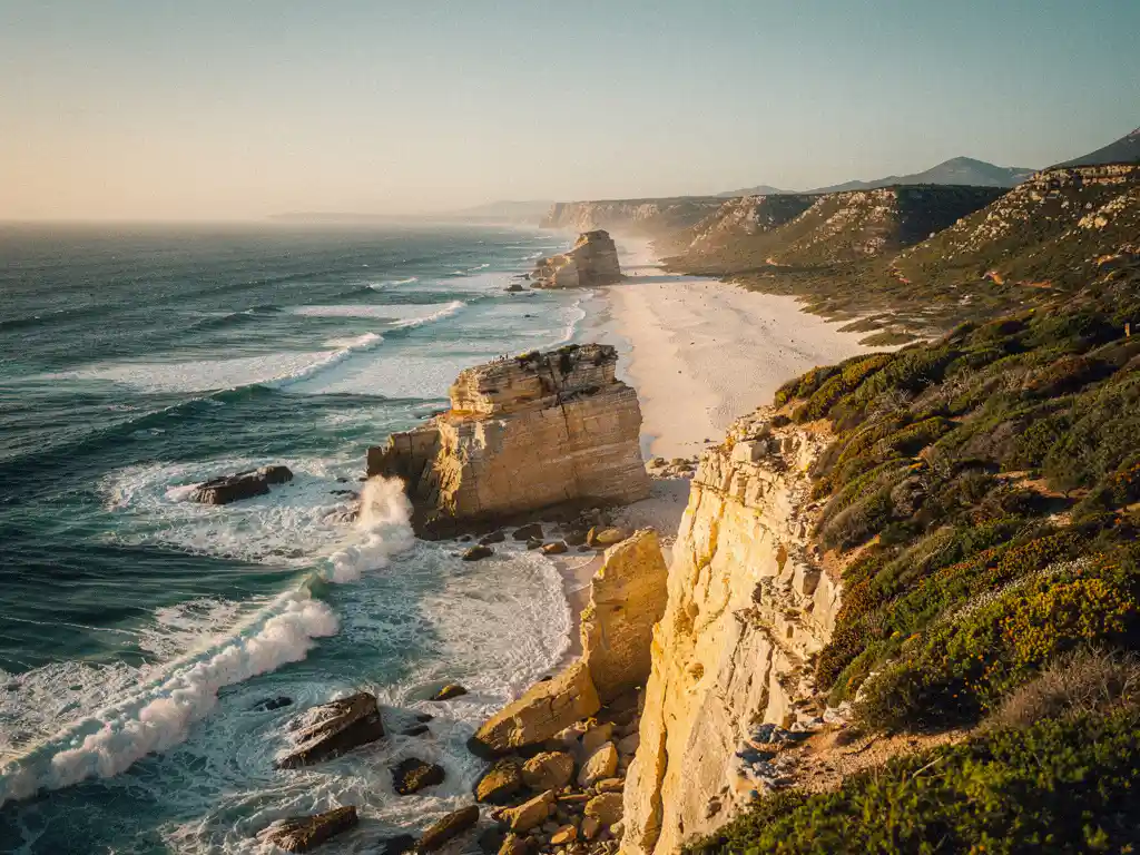 Rotsachtige kustlijn van Zuid-Afrika met turquoise golven tegen gouden zandstenen kliffen, wit strand en fynbos-vegetatie in middagzon.