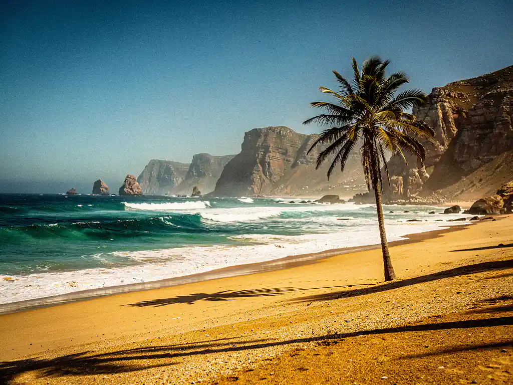 Zonnig Zuid-Afrikaans strand in februari met goudkleurig zand, turquoise golven en dramatische rotsklif onder strakblauwe hemel.