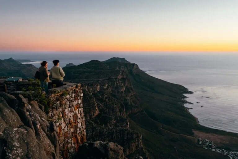 Twee vrouwelijke reizigers kijken vanaf de top van de Tafelberg hoe de zon in zee zakt