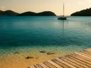 Verouderd houten steiger boven turquoise Caribisch water met drie tropische eilanden aan de horizon en een zeilboot bij zonsondergang.