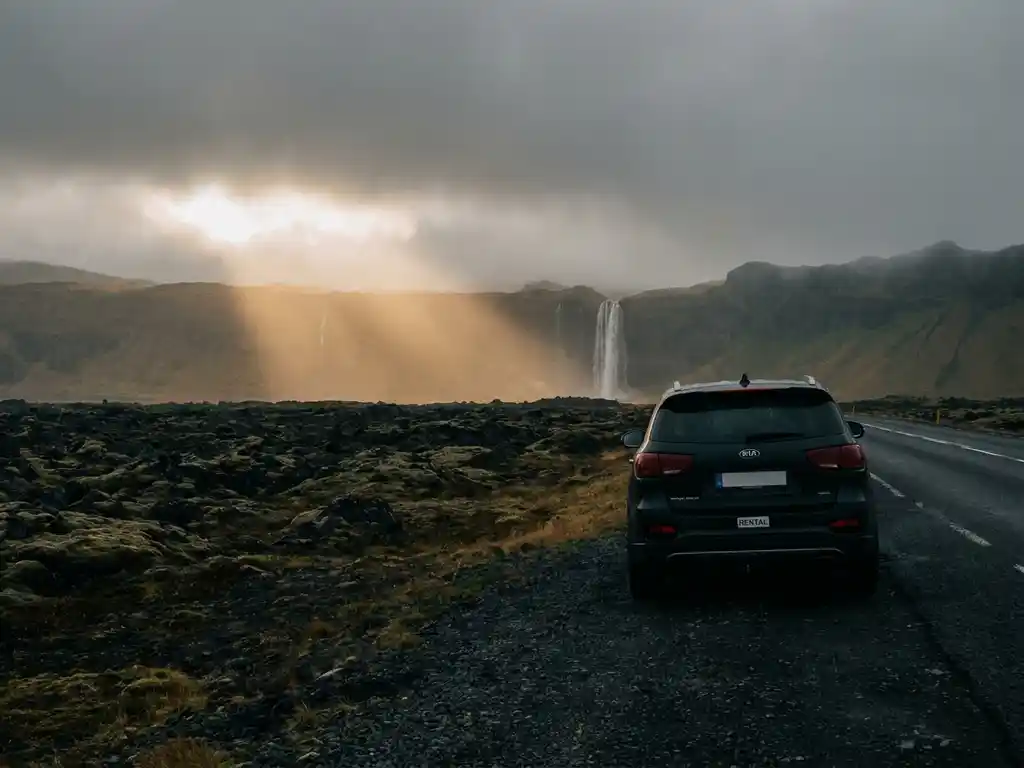 Huurauto geparkeerd langs IJslandse ringweg, met zwarte lavavelden, een waterval in de verte en een gouden lichtstraal door bewolkte lucht.