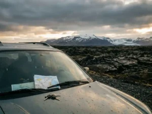 Versleten huurauto op IJslands Ring Road met zwarte lavavelden en besneeuwde vulkaan, wegenkaart en autosleutels op dashboard.