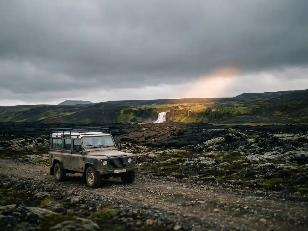 Terreinwagen op IJslands hooglandweg, omgeven door zwarte lavavelden, mosrijke kliffen met waterval en dramatisch goudkleurig licht.