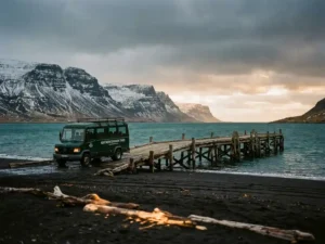 Verweerde houten steiger in een IJslandse fjord met zwart lavastrand, besneeuwde basaltrotsen en een minibus op de oever.