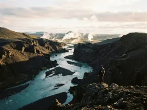 Eenzame wandelaar kijkt uit over IJslands vulkanisch landschap met turquoise gletsjerivier en opstijgende geothermische stoom bij gouden uur.